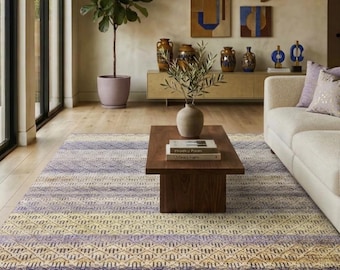 May include: A rectangular area rug featuring a repeating geometric pattern in shades of muted yellow, lavender, and blue. The rug has a textured weave and a short fringe along the edges, covering a light-coloured wooden floor in a living room space.