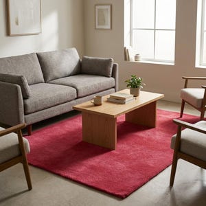 May include: A rectangular area rug in a solid deep red color with a plush texture, positioned beneath a light wood coffee table, a gray sofa, and two light-colored armchairs with wooden frames in a bright, neutral-toned living room space.