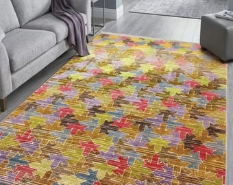 May include: A rectangular area rug featuring a repeating pattern of stylised maple leaves in shades of yellow, red, purple, blue, and green. The rug has a textured surface with horizontal lines and short white fringe along the two shorter edges.