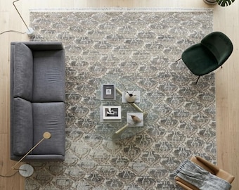 May include: A large rectangular area rug featuring a distressed, faded floral pattern in shades of beige, grey, and muted blue. The rug has a short fringe on the top and bottom edges and is positioned under a grey sofa, a glass coffee table, and chairs.