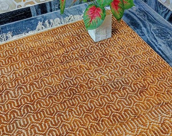 May include: A rectangular area rug featuring a repeating golden-brown and white geometric wave pattern with a textured surface and white fringe along the edge. A potted plant with large green and red heart-shaped leaves sits on the rug.