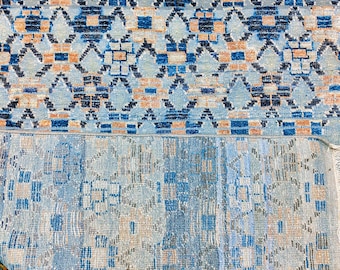 May include: A woven rug featuring a repeating geometric pattern in shades of light blue, dark blue, and peach. The top section displays a crisp, detailed design, while the bottom section shows the reverse side with a muted, textured appearance.