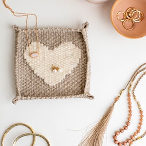 May include: A woven heart-shaped jewelry holder with a beige and white color scheme. The holder is filled with a gold necklace, a pair of pearl earrings, and a beaded necklace with a tassel.