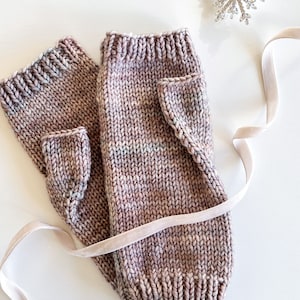 May include: A pair of knitted fingerless gloves in a pink and white color scheme with a speckled pattern. The gloves are folded and tied together with a pink ribbon.
