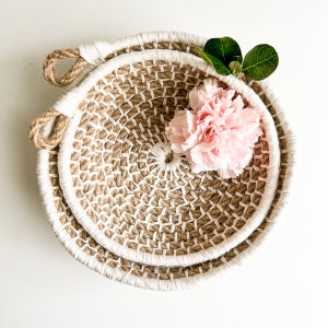 May include: Two round woven baskets made of natural jute rope with a white trim. The baskets are nested together and a pink flower is placed in the center of the top basket.