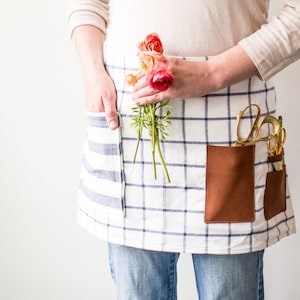 May include: A white and blue checkered apron with a brown leather pocket and a pair of gold scissors. The apron has a striped pocket on the left side and a person is holding a bouquet of pink and orange flowers.