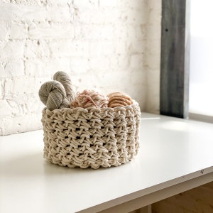 May include: A beige macrame basket filled with balls of yarn in shades of pink, peach, and gray. The basket is sitting on a white table.