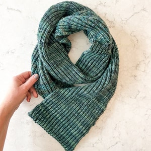 May include: A green and blue knitted scarf with a ribbed texture. The scarf is folded in half and laid flat on a white surface.