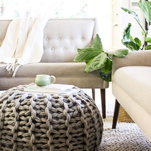 May include: A large gray knitted ottoman with a chunky knit pattern sits on a light beige rug in a living room. A white couch with a throw blanket and a green plant are in the background.