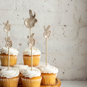 May include: A tiered cake stand with six cupcakes topped with white frosting and decorated with crocheted bunny rabbit toppers. The cupcakes are on a wooden cake stand with a white background.
