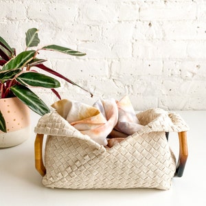 May include: A woven basket with wooden handles, filled with colorful fabric scraps. The basket is beige and has a woven pattern. The fabric scraps are in shades of pink, orange, and yellow.