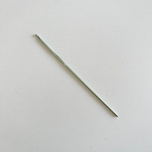 May include: A silver crochet hook, angled diagonally across a white surface. The hook has a rounded head on one end and a straight shaft. The hook is a crafting tool used for creating crocheted items.