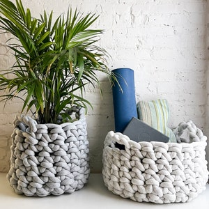 May include: Two gray, chunky knit baskets. One holds a green plant, the other a blue yoga mat, a gray yoga block, and a striped bolster. The baskets are on a white surface against a white brick wall.