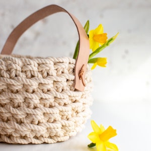 May include: A white crocheted basket with a brown leather handle. The basket is filled with yellow daffodils.