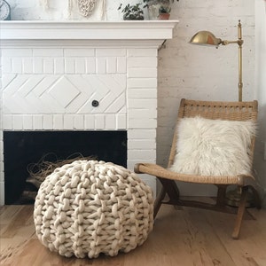 May include: A cozy living room scene featuring a cream-colored, woven pouf, a wooden chair with a fluffy white pillow, and a brass lamp. A white brick fireplace is in the background, creating a warm and inviting atmosphere.