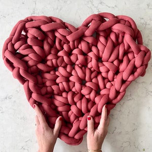 May include: A large, heart-shaped knitted pillow in a deep rose color. The pillow is made with thick yarn and has a chunky, textured look.