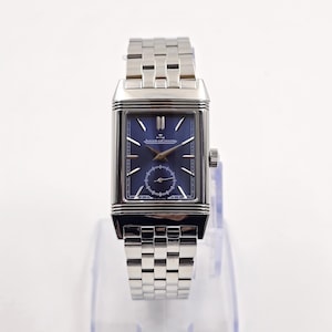 May include: A rectangular stainless steel wristwatch with a blue dial. The watch features a silver-tone bracelet and a small seconds subdial. The watch has the text "JAEGER-LECOULTRE" on the dial.