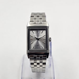 May include: A silver rectangular wristwatch with a silver dial and a stainless steel band. The watch features a small seconds subdial and the brand name "Jaeger-LeCoultre" on the dial. The watch is displayed on a clear stand.