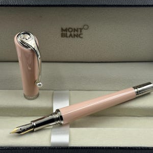 Puede incluir: Una pluma estilográfica Montblanc rosa con detalles plateados, presentada en una caja negra. La pluma tiene un clip plateado y un plumín dorado. El logotipo de Montblanc es visible en el capuchón. Instrumento de escritura de lujo.