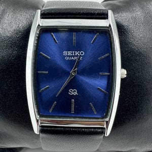 Könnte beinhalten: Eine rechteckige Seiko Quartz-Uhr mit tiefblauem Zifferblatt und silberfarbenem Gehäuse. Die Uhr hat ein schwarzes Lederarmband und wird auf einer schwarzen Oberfläche präsentiert. Das Zifferblatt zeigt das Seiko-Logo und das Wort "Quartz".