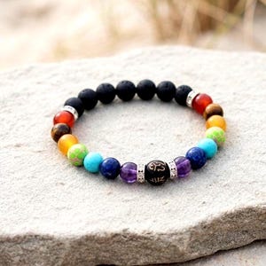 Puede incluir: Una pulsera elástica de cuentas con una serie de piedras redondas multicolores que incluyen morado, azul, turquesa, verde, amarillo, naranja y rojo, acentuadas por cuentas de lava negra y una cuenta central negra con símbolos blancos grabados.