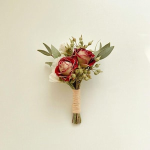 Pode incluir: Um pequeno boutonniere floral com duas rosas vermelhas e centros creme, rodeado por folhas verdes e pequenos bot&otilde;es verdes. As hastes s&atilde;o enroladas em barbante bege, criando um visual r&uacute;stico e elegante. Perfeito para casamentos ou eventos.