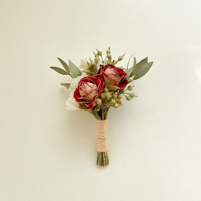 Pode incluir: Um pequeno boutonniere floral com duas rosas vermelhas e centros creme, rodeado por folhas verdes e pequenos bot&otilde;es verdes. As hastes s&atilde;o enroladas em barbante bege, criando um visual r&uacute;stico e elegante. Perfeito para casamentos ou eventos.