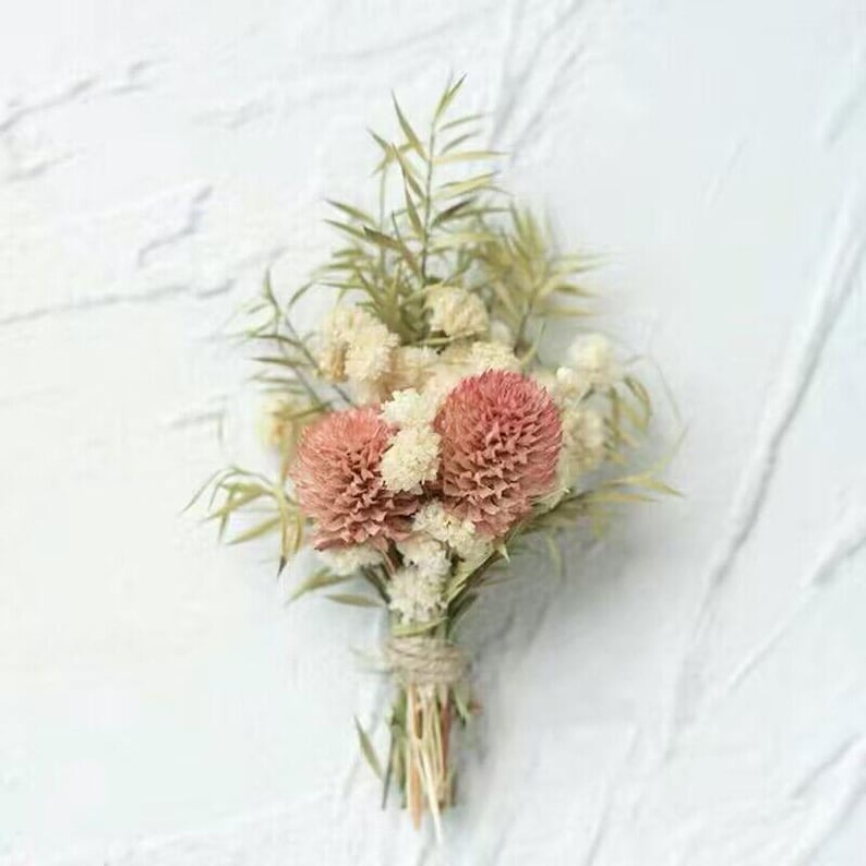 Pode incluir: Um pequeno buqu&ecirc; de flores secas, com duas flores rosas redondas proeminentes. O arranjo inclui folhagem branca e verde delicada, amarrada com barbante. O fundo &eacute; uma superf&iacute;cie branca texturizada.