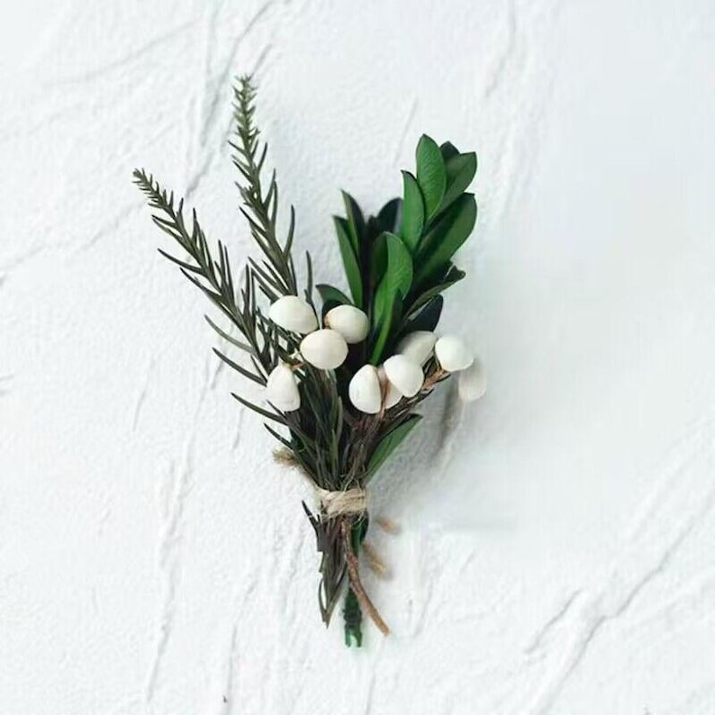 Pode incluir: Um pequeno buqu&ecirc; de vegeta&ccedil;&atilde;o e bagas brancas amarrado com barbante. O arranjo apresenta galhos verdes escuros, folhas verdes brilhantes e pequenas bagas brancas. O fundo &eacute; uma superf&iacute;cie branca texturizada.