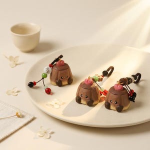 May include: Three small, brown wooden charms shaped like pudding cups, each with a pink top and a smiling face. They are adorned with beaded strings in red, green, and black. The charms sit on a white plate, next to a small teacup.