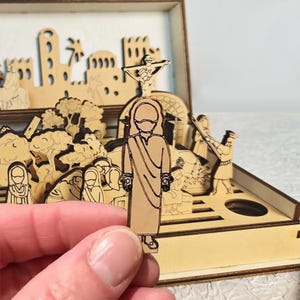 K&ouml;nnte beinhalten: Ein Holz-Diorama, das eine religi&ouml;se Szene darstellt, mit Figuren, Geb&auml;uden und einem Kreuz. Das Diorama besteht aus hellem Holz mit aufwendigen Ausschnitten. Eine Figur wird vor dem Diorama gehalten.