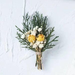 Pode incluir: Um pequeno boutonniere artesanal com rosas amarelas secas, gipsofila branca e raminhos verdes perenes. Os caules s&atilde;o amarrados com barbante, criando uma est&eacute;tica r&uacute;stica e natural. Perfeito para casamentos ou ocasi&otilde;es especiais.