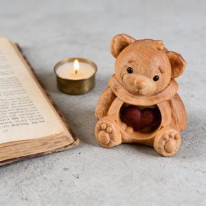 May include: A wooden teddy bear figurine with a carved heart in its chest. The bear is light brown with dark eyes and a carved scarf. A lit candle in a brass holder and an open book are in the background.