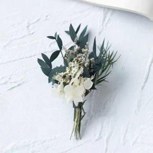 Pode incluir: Um pequeno buqu&ecirc; de flores brancas e verdes, incluindo hort&ecirc;nsias brancas e folhas de eucalipto verde escuro, amarrado com uma fita verde. O arranjo &eacute; colocado sobre um fundo branco texturizado, criando uma est&eacute;tica natural e elegante.