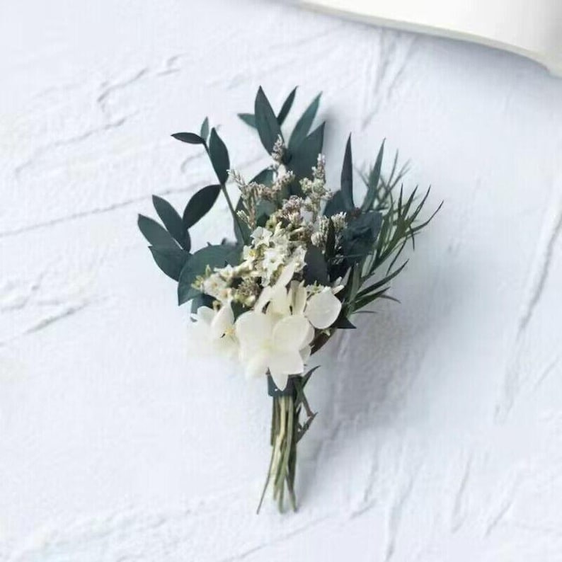Pode incluir: Um pequeno buqu&ecirc; de flores brancas e verdes, incluindo hort&ecirc;nsias brancas e folhas de eucalipto verde escuro, amarrado com uma fita verde. O arranjo &eacute; colocado sobre um fundo branco texturizado, criando uma est&eacute;tica natural e elegante.