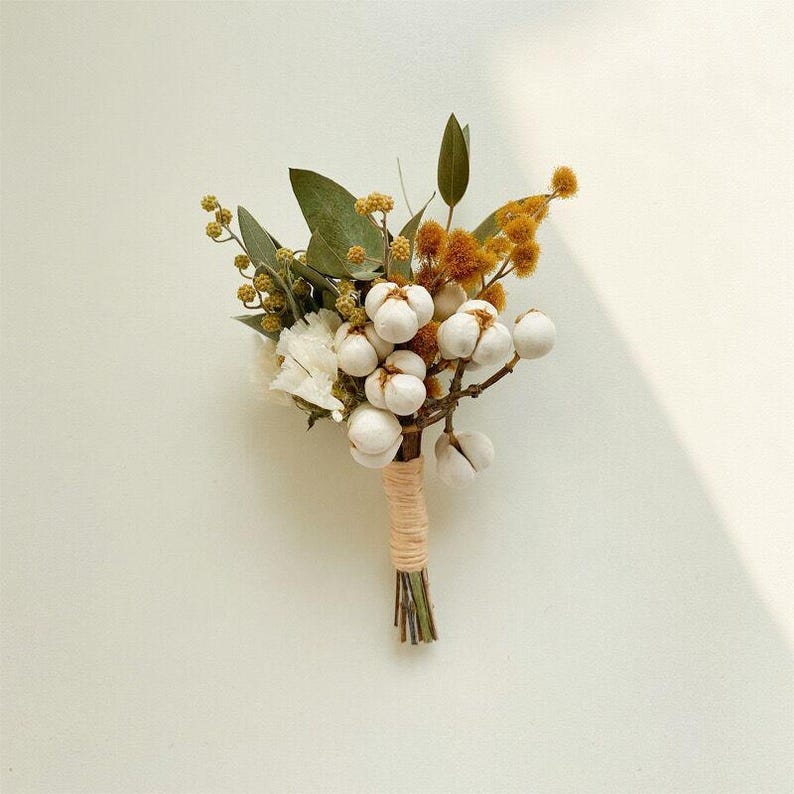 Pode incluir: Uma pequena boutonniere artesanal com bot&otilde;es de algod&atilde;o branco, flores secas amarelas e castanhas e folhas verdes. As hastes s&atilde;o enroladas com barbante bege, criando uma est&eacute;tica r&uacute;stica e natural. Perfeito para casamentos ou eventos especiais.