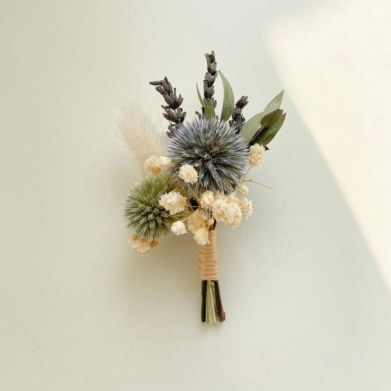 Pode incluir: Um pequeno arranjo floral seco com um cardo azul, cardo globo verde, lavanda e pequenas flores brancas. As hastes s&atilde;o amarradas com barbante. Um raminho de capim-dos-pampas branco e fofo adiciona textura. Perfeito para um casamento.