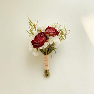 Pode incluir: Um pequeno buqu&ecirc; com duas rosas vermelhas escuras e cravos brancos, acentuado com folhagem verde. As hastes s&atilde;o embrulhadas com barbante bege, criando um arranjo floral r&uacute;stico e elegante.
