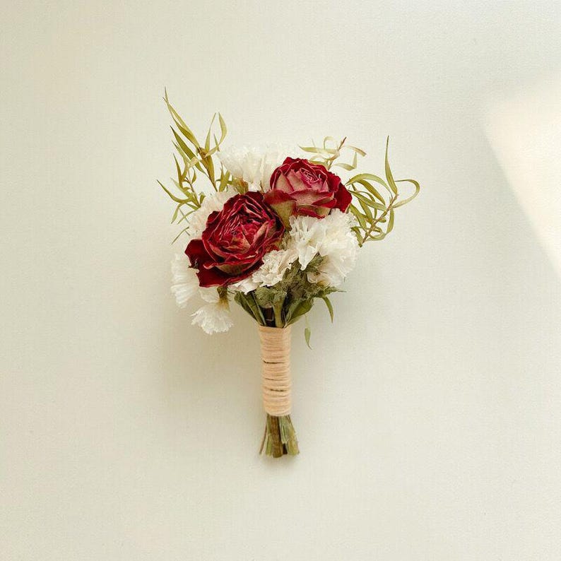Pode incluir: Um pequeno buqu&ecirc; com duas rosas vermelhas escuras e cravos brancos, acentuado com folhagem verde. As hastes s&atilde;o embrulhadas com barbante bege, criando um arranjo floral r&uacute;stico e elegante.