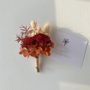 Pode incluir: Uma boutonniere floral bord&ocirc; e laranja com flores secas e vegeta&ccedil;&atilde;o, amarrada com barbante. Um cart&atilde;o branco com uma ilustra&ccedil;&atilde;o de flor e as palavras "THANK YOU" est&aacute; ao lado da boutonniere.