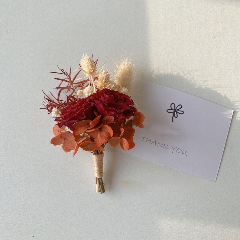 Pode incluir: Uma boutonniere floral bord&ocirc; e laranja com flores secas e vegeta&ccedil;&atilde;o, amarrada com barbante. Um cart&atilde;o branco com uma ilustra&ccedil;&atilde;o de flor e as palavras "THANK YOU" est&aacute; ao lado da boutonniere.