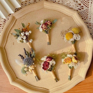 Pode incluir: Uma cole&ccedil;&atilde;o de cinco pequenos buqu&ecirc;s de flores secas dispostos em uma bandeja bege com bordas onduladas. Cada buqu&ecirc; apresenta uma variedade de flores em tons de branco, amarelo e vermelho, amarradas com barbante. Os buqu&ecirc;s s&atilde;o um arranjo floral r&uacute;stico e charmoso.