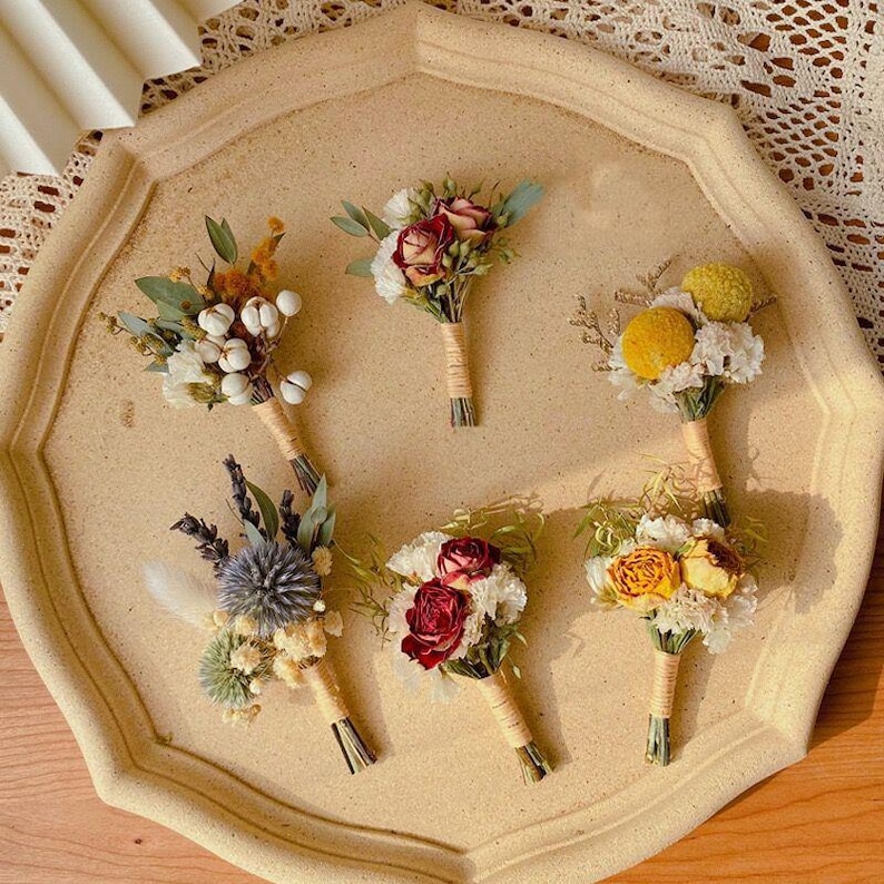Pode incluir: Uma cole&ccedil;&atilde;o de cinco pequenos buqu&ecirc;s de flores secas dispostos em uma bandeja bege com bordas onduladas. Cada buqu&ecirc; apresenta uma variedade de flores em tons de branco, amarelo e vermelho, amarradas com barbante. Os buqu&ecirc;s s&atilde;o um arranjo floral r&uacute;stico e charmoso.