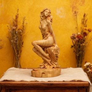 May include: A wooden sculpture of a seated woman with flowing hair and draped clothing. The figure is carved from light-colored wood and sits on a textured base. The sculpture is displayed on a wooden table with dried flowers in the background.