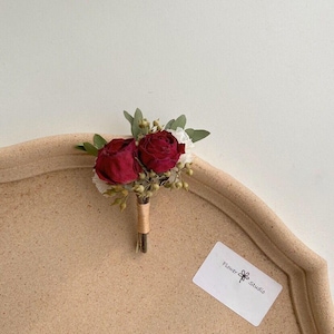 Pode incluir: Uma pequena boutonniere floral com duas rosas vermelhas escuras e flores brancas, acentuada com folhas verdes e pequenas bagas. Os caules s&atilde;o embrulhados em barbante. Um pequeno cart&atilde;o branco com o texto "Flower Studio" &eacute; vis&iacute;vel.