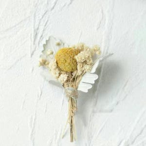 Pode incluir: Um pequeno buqu&ecirc; de flores secas com uma grande flor esf&eacute;rica amarela rodeada por flores menores creme. O buqu&ecirc; &eacute; amarrado com barbante e inclui detalhes brancos em forma de p&eacute;tala. O fundo &eacute; uma superf&iacute;cie branca texturizada.
