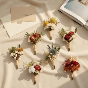 May include: An assortment of seven small, dried flower bouquets with stems wrapped in twine. The bouquets feature a variety of colors, including red, yellow, white, and green. A cream-colored envelope with a wax seal and a book are in the background.