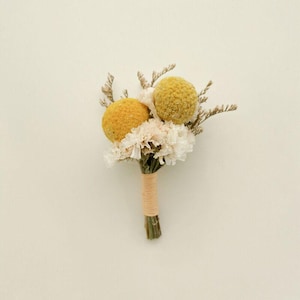 Pode incluir: Uma pequena boutonniere artesanal com dois billy buttons esf&eacute;ricos amarelos, flores brancas e rosa p&aacute;lido e raminhos secos. As hastes s&atilde;o enroladas com barbante bege, criando um arranjo floral r&uacute;stico e elegante.