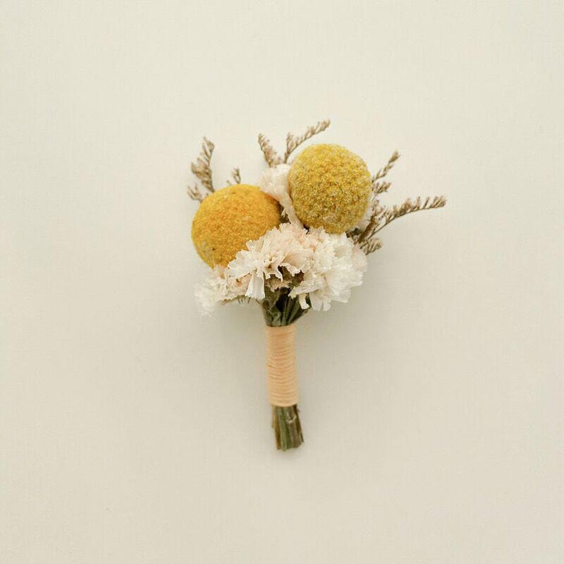 Pode incluir: Uma pequena boutonniere artesanal com dois billy buttons esf&eacute;ricos amarelos, flores brancas e rosa p&aacute;lido e raminhos secos. As hastes s&atilde;o enroladas com barbante bege, criando um arranjo floral r&uacute;stico e elegante.