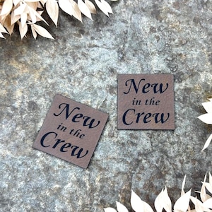 Op de afbeelding: Twee vierkante, bruine labels met de tekst "New in the Crew" in zwart schrift worden weergegeven op een getextureerd, grijs oppervlak. De labels zijn omgeven door gedroogd, crèmekleurig gebladerte.